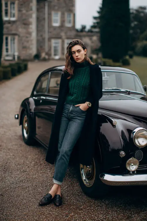 A woman wears high-rise jeans with a forest green cable-knit sweater, topped with a black wool coat and loafers, full body near a vintage car parked outside a winter estate