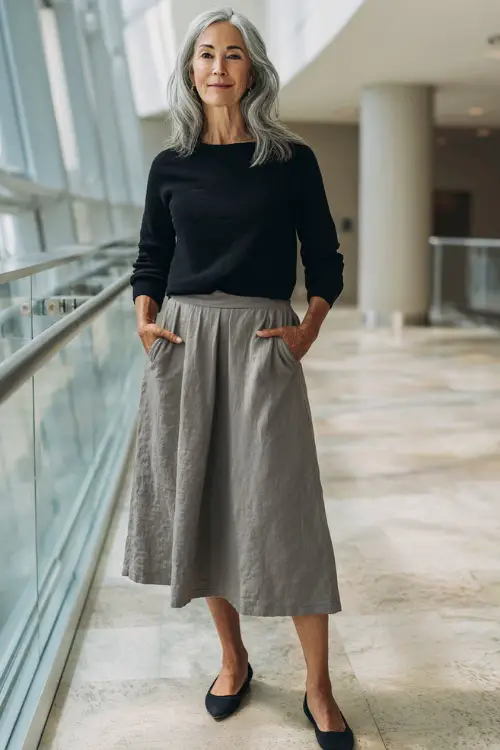 Full-body view of a woman over 40 wearing a plain midi skirt with a tucked-in knit top and simple flats, understated casual business casual look, modern indoor office environment 
