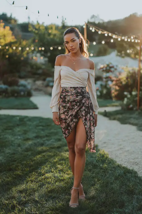 A chic woman in a floral wrap skirt, off-shoulder blouse, heeled sandals, delicate jewelry, outdoor evening date setting with string lights, full-body portrait