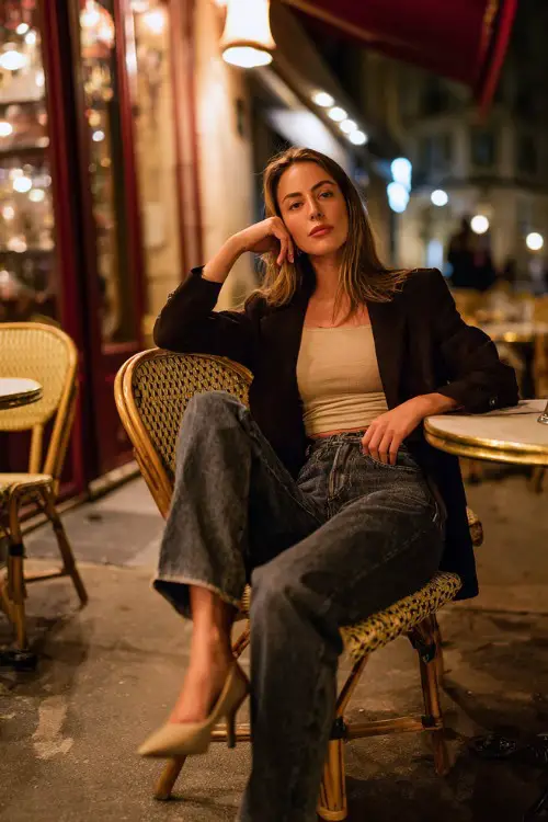 A chic woman in dark straight-leg jeans, fitted knit top, cropped blazer, pointed heels, cozy café evening background suggesting a relaxed night out with friends, full-body fashion photography 