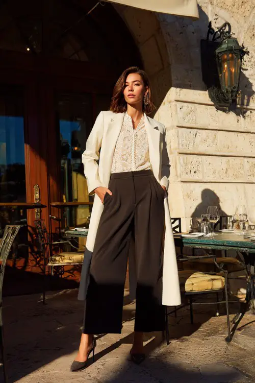 A chic woman in wide-leg trousers, lace blouse, structured long coat, high heels, upscale restaurant exterior background, full-body editorial fashion photography 