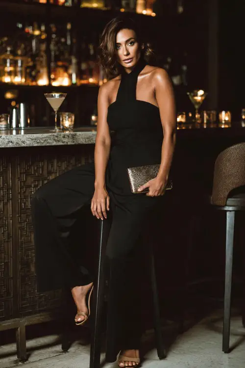 A chic woman wearing a sleek black jumpsuit with high heels and a metallic clutch, posing next to a bar stool in a luxury cocktail lounge