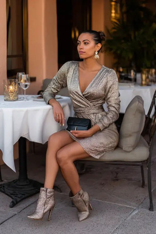 A fashionable woman wearing a mini wrap dress with heeled ankle boots, statement earrings, sleek clutch, romantic restaurant patio background, full-body shot