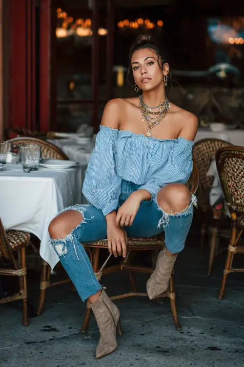 A fashionable woman wearing ripped boyfriend jeans, off-shoulder blouse, heeled ankle boots, layered necklaces, casual restaurant patio background