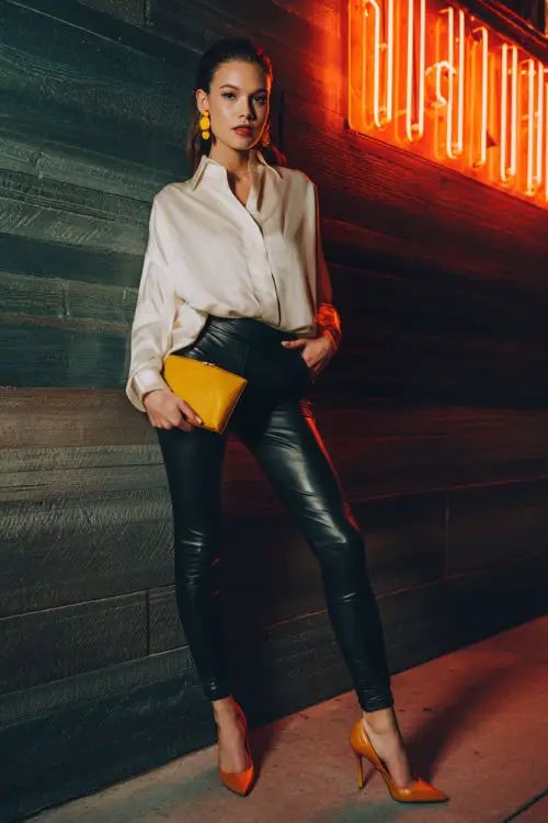 A stylish woman wearing leather leggings with an oversized blouse slightly tucked in and pointed heels, bold earrings and a sleek clutch, posing near a neon-lit bar wall, full-body composition 