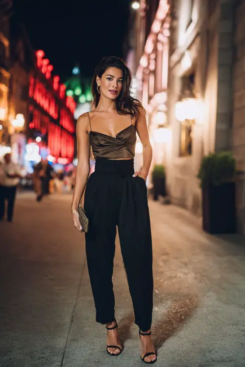 A stylish woman wearing tailored high-waisted trousers, silky camisole top, strappy heels, elegant clutch bag, city dinner street background at night, full-body fashion photography, cinematic lighting