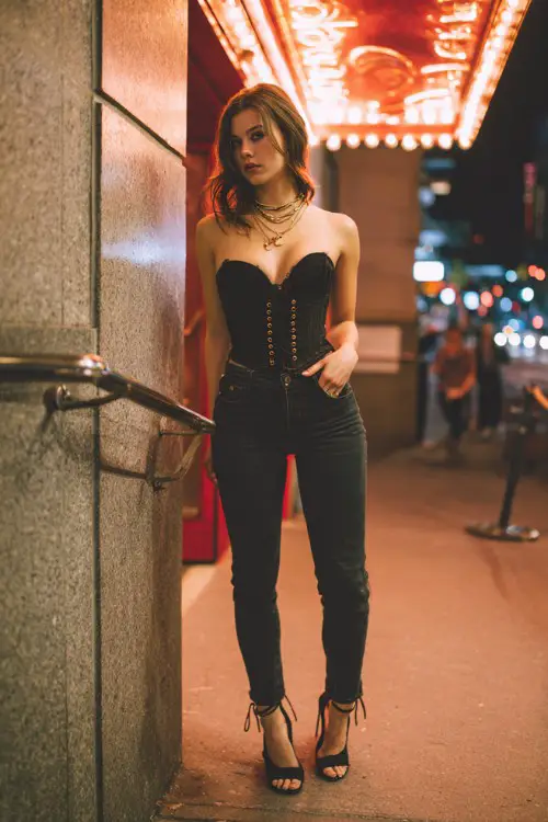 A trendy woman wearing dark skinny jeans with a fitted corset top and strappy heels, accessorized with layered necklaces, standing outside a trendy night bar entrance under glowing signage