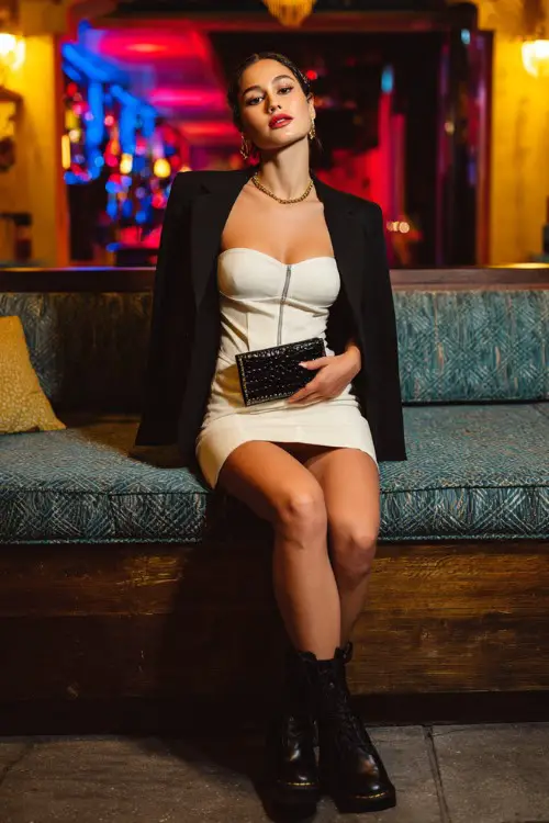 A woman in a bodycon mini dress, chunky platform boots, cropped blazer, bold clutch, trendy cocktail bar interior background, full-body fashion photo