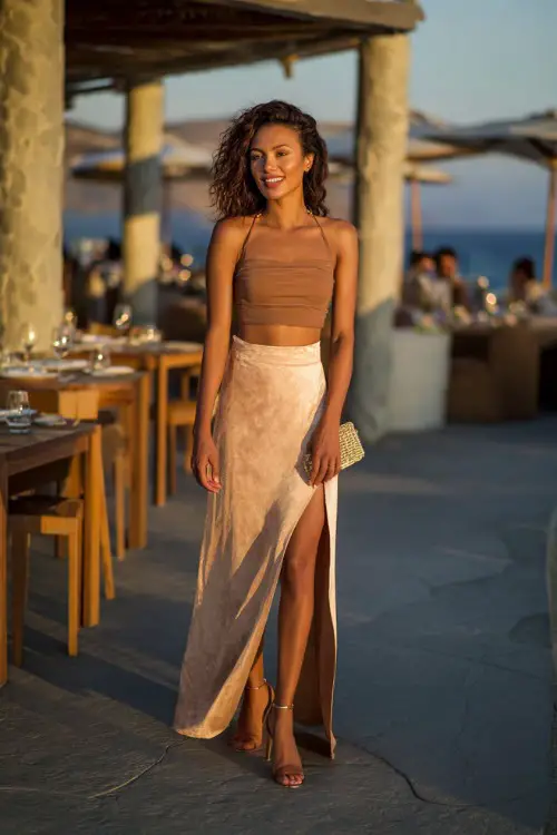 A woman wearing a high-slit maxi skirt, fitted crop top, heeled sandals, chic clutch, sunset beachside restaurant background, full-body fashion photography