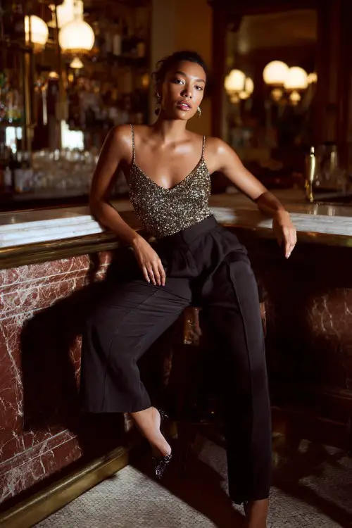 A woman wearing high-waisted tailored trousers with a sparkly sequin top and sleek stilettos, posing inside an upscale lounge bar with dim lighting and marble bar counter, full-body portrait 