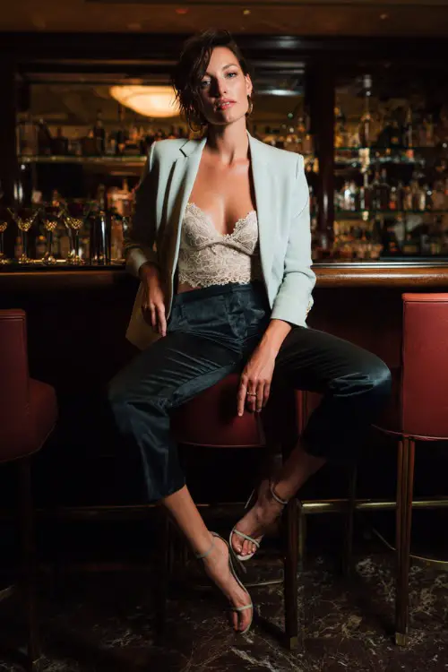 A woman wearing slim ankle-length trousers, lace camisole, cropped blazer, heeled sandals, cocktail bar interior background, full-body portrait, dramatic evening lighting, fashion photography style 