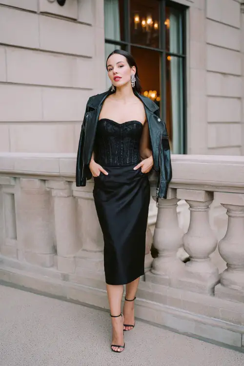 A woman wears a classy rocker cocktail guest dress in black satin with a structured corset bodice and subtle lace overlay, styled with sleek heels, a cropped leather jacket and statement earrings