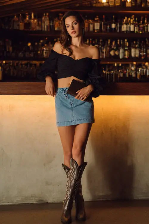 A chic woman wearing a high-waisted denim mini skirt with a fitted off-shoulder blouse and tall cowgirl boots,