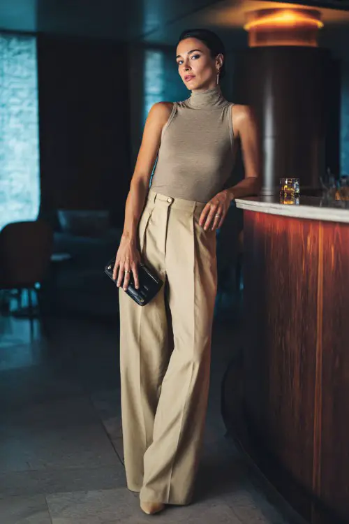 A chic woman wearing tailored wide-leg trousers with a sleeveless fitted top and block heels, holding a sleek clutch, posing confidently inside a modern lounge bar with moody lighting 