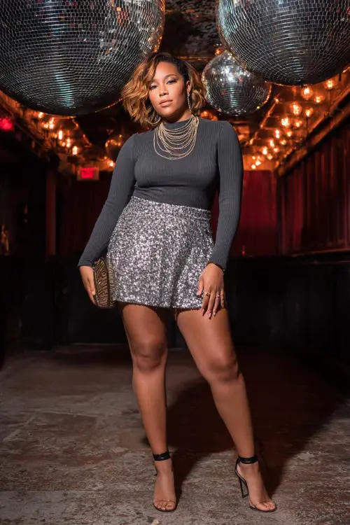 A confident plus-size woman wearing a sparkly sequin mini skirt with a fitted long-sleeve top and ankle-strap heels, accessorized with layered necklaces and a clutch