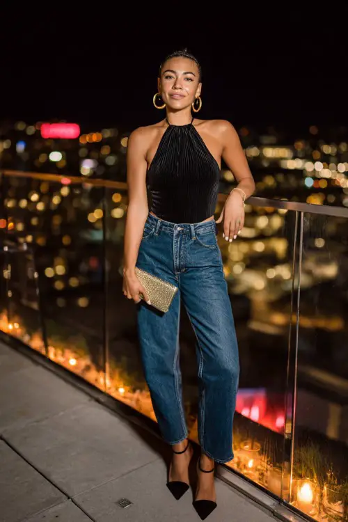 A confident woman wearing high-waisted straight-leg blue jeans with a sleek black halter top and strappy high heels, accessorized with bold hoop earrings and a small metallic clutch