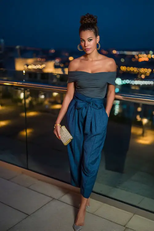 A confident woman wearing high-waisted tailored trousers with a fitted off-shoulder top and sleek heels, accessorized with hoop earrings and a clutch