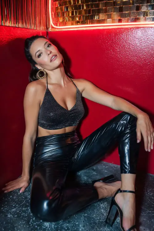 A confident woman wearing shiny faux-leather leggings with a cropped sparkly top and strappy stilettos, accessorized with bold hoop earrings, posing inside a neon-lit nightclub bar, full-body frame 