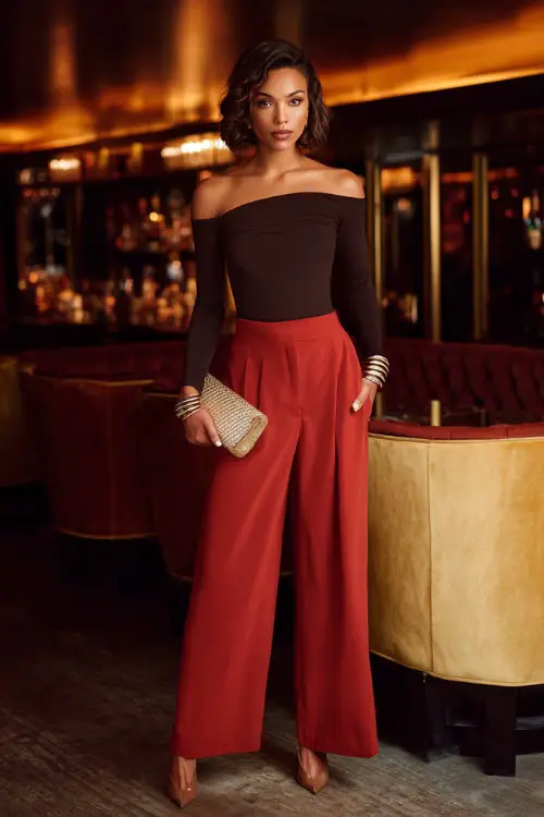A confident woman wearing tailored wide-leg trousers with a fitted long-sleeve off-shoulder top and sleek heels, accessorized with layered bracelets and a structured clutch 