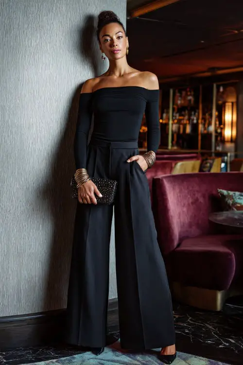 A confident woman wearing tailored wide-leg trousers with a fitted long-sleeve off-shoulder top and sleek heels, accessorized with layered bracelets and a structured clutch