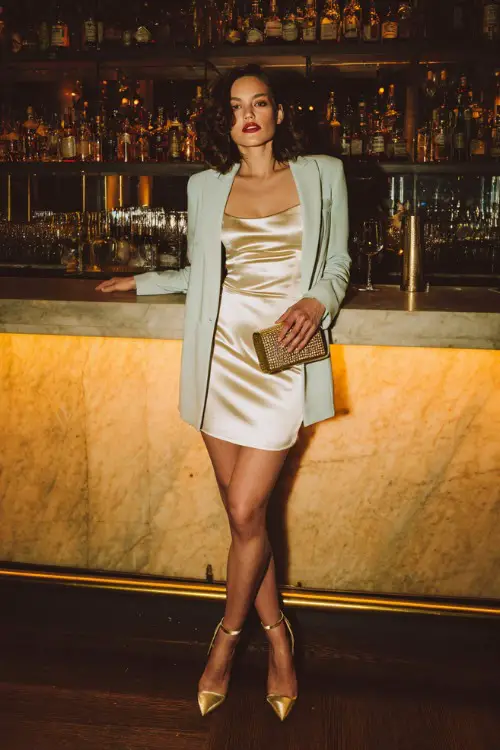 A fashionable woman wearing a satin slip dress layered with a lightweight long-sleeve blazer and strappy heels