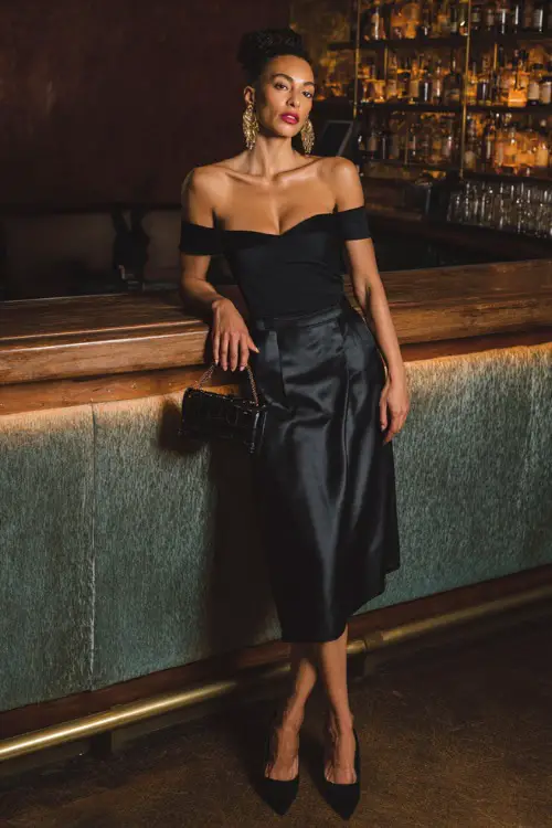 A fashionable woman wearing a sleek black satin midi skirt with a fitted off-shoulder blouse and pointed-toe heels, accessorized with statement earrings and a mini handbag