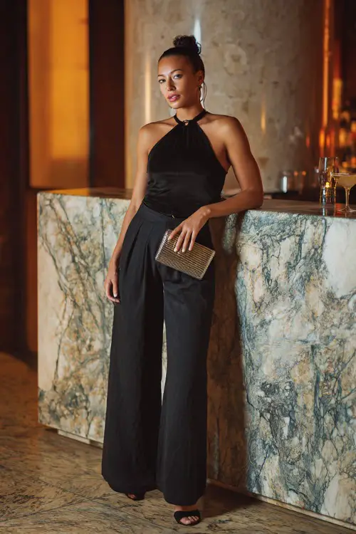 A fashionable woman wearing black wide-leg trousers with a fitted halter top and strappy heels, holding a metallic clutch while posing near a marble cocktail bar counter