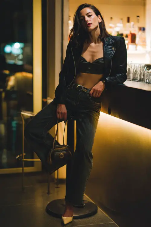 A fashionable woman wearing high-waisted dark jeans with a fitted crop top and a structured leather jacket, paired with pointed-toe heels and a mini shoulder bag