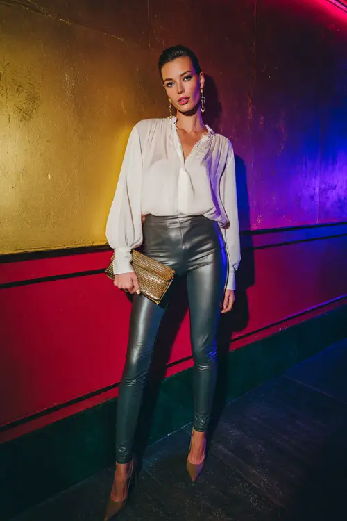 A fashionable woman wearing leather leggings with an oversized blouse slightly tucked in and pointed heels, bold earrings and a sleek clutch, standing near a neon-lit wall inside a trendy night bar