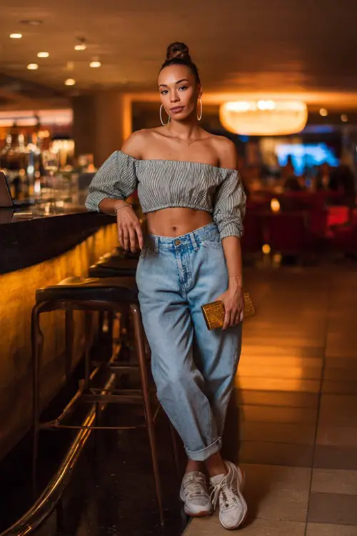 A fashionable woman wearing relaxed denim jeans with a fitted off-shoulder blouse and trendy sneakers, accessorized with hoop earrings and a sleek clutch