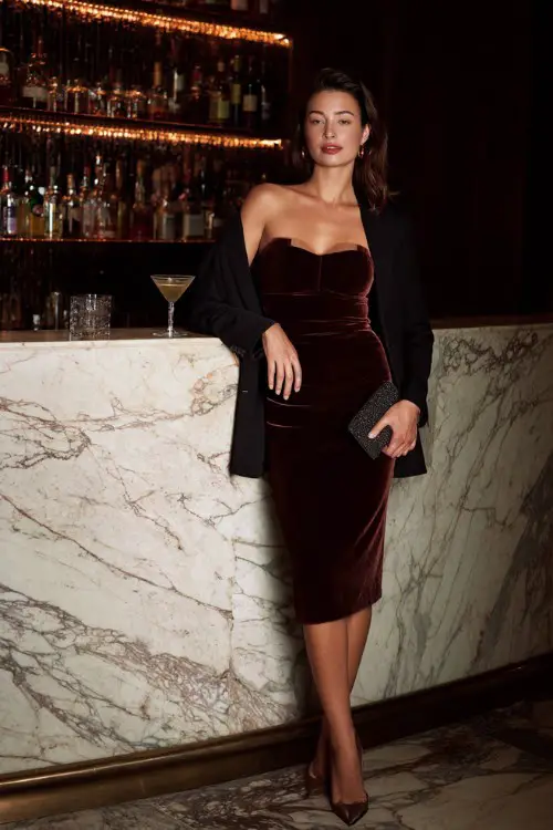 A glamorous woman wearing a velvet midi dress layered with a structured blazer and elegant heels, holding a clutch while standing beside a marble cocktail bar counter, full-body composition