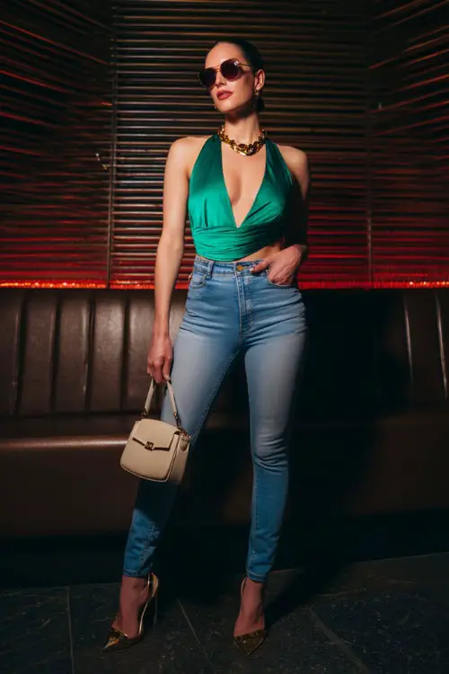 A glamorous woman wearing high-waisted skinny jeans with a bold asymmetrical top and pointed heels, accessorized with layered gold jewelry and a mini handbag 