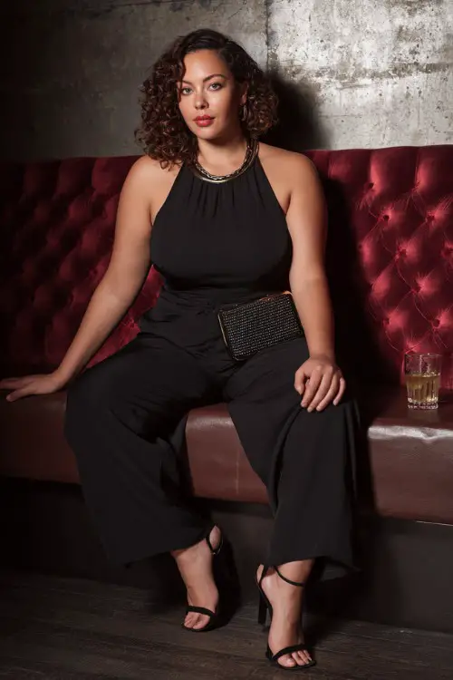 A stylish plus-size woman wearing a sleek black jumpsuit with high heels and a structured mini handbag, soft curls hairstyle and statement jewelry 