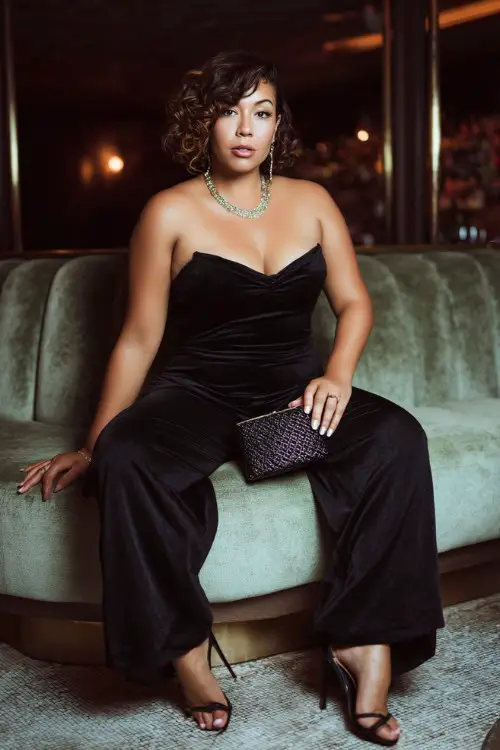 A stylish plus-size woman wearing a sleek black jumpsuit with high heels and a structured mini handbag, soft curls hairstyle and statement jewelry