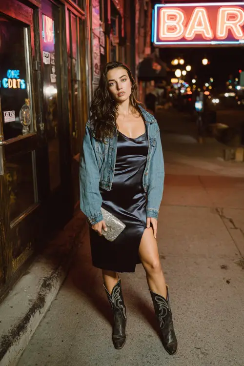 A stylish woman wearing a satin slip dress layered with a cropped denim jacket and cowgirl boots