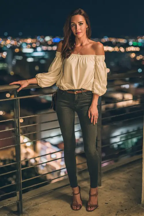A stylish woman wearing dark skinny jeans with an off-shoulder blouse and heeled sandals, delicate jewelry and a mini shoulder bag, standing near a rooftop bar railing with city lights behind her 