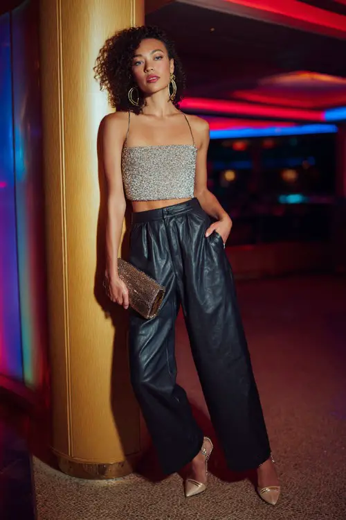 A stylish woman wearing high-waisted leather pants with a sparkly cropped top and pointed heels, accessorized with hoop earrings and a metallic clutch, posing under colorful LED club lights