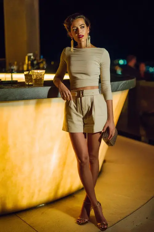 A stylish woman wearing high-waisted tailored shorts with a fitted long-sleeve top and sleek heels, accessorized with statement earrings and a mini handbag