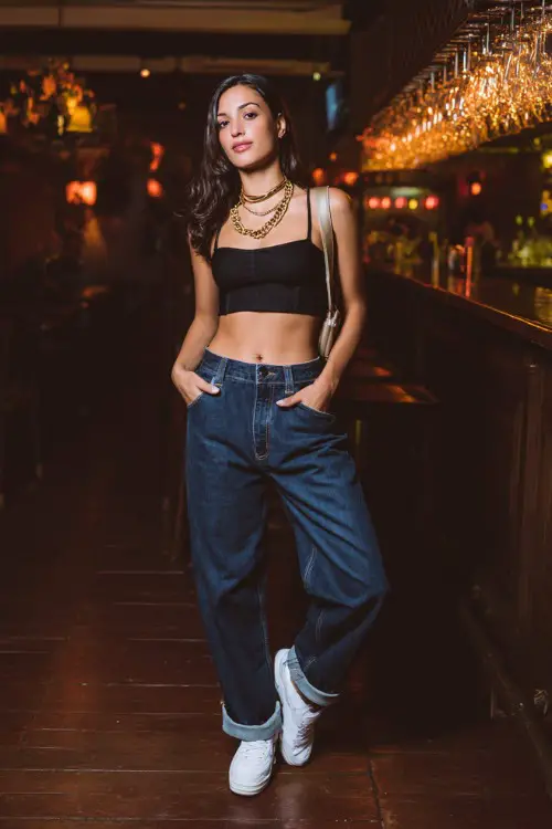 A stylish woman wearing straight-leg blue jeans with a fitted black crop top and clean white sneakers, layered gold necklaces and a small shoulder bag