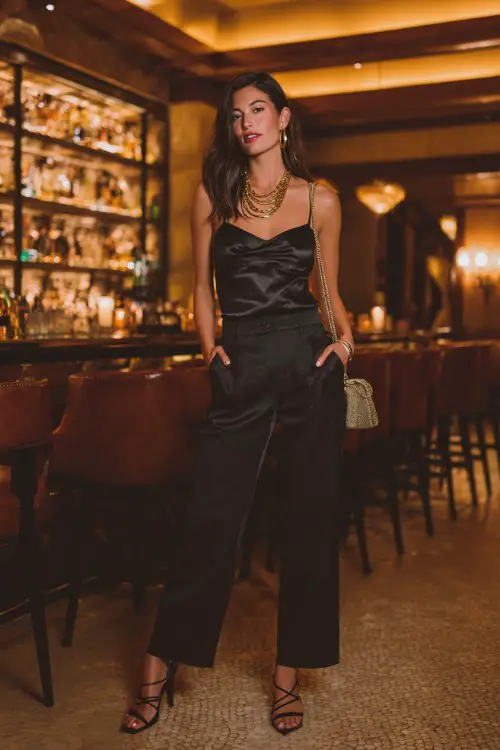 A stylish woman wearing straight-leg dark jeans with a fitted black satin cami and strappy heels, layered gold necklaces and a small shoulder bag 