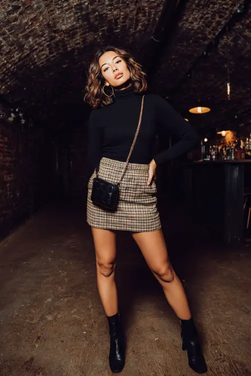 A trendy woman wearing a plaid mini skirt with a fitted black turtleneck top and ankle boots, accessorized with a crossbody bag and hoop earrings 