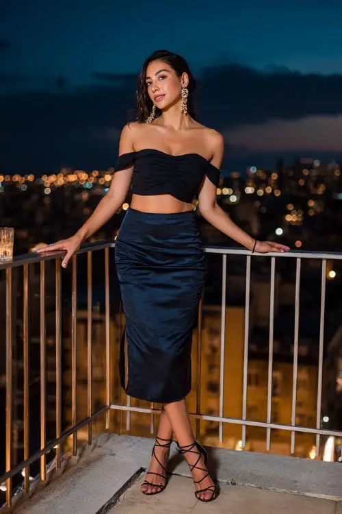 A trendy woman wearing a satin midi skirt with a fitted off-shoulder top and strappy heels, accessorized with statement earrings and a mini handbag