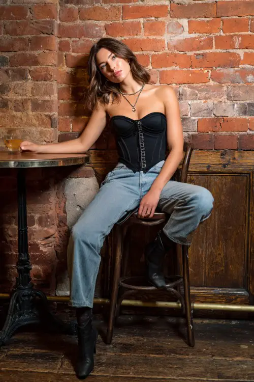 A trendy woman wearing straight-leg jeans with a fitted corset-style top and ankle boots, accessorized with layered necklaces and a small handbag