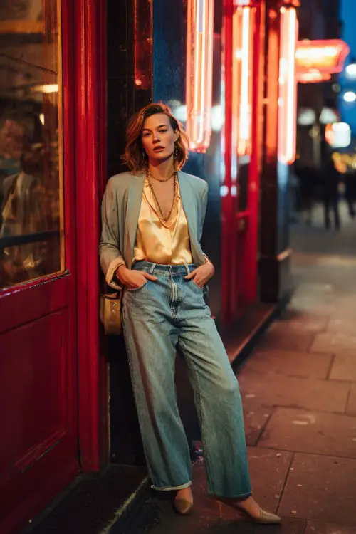 A trendy woman wearing straight-leg jeans with a silky blouse and a lightweight tailored jacket, accessorized with layered jewelry and a mini handbag 