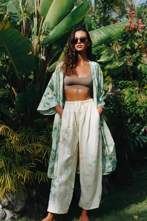 A woman wears a simple tank top layered with a lightweight kimono and flowy wide-leg pants, styled with flat sandals and oversized sunglasses