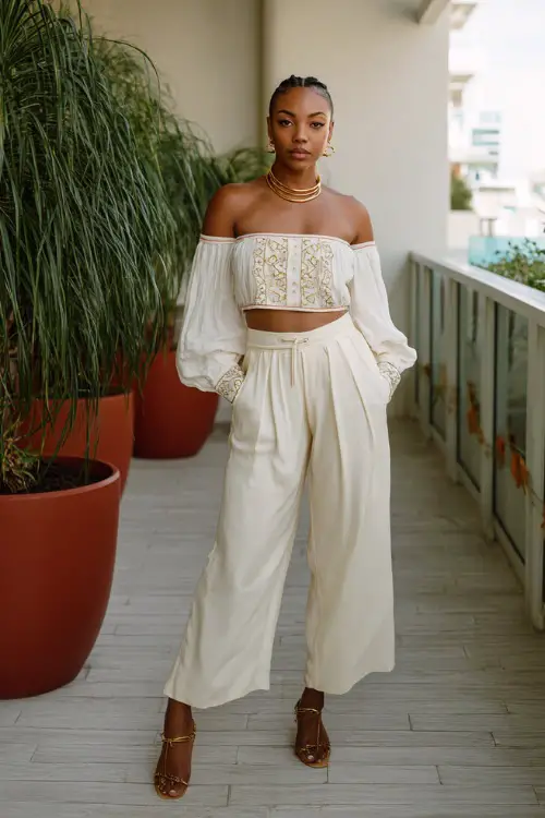 A Black woman wears a lightweight white off-shoulder blouse with embroidery paired with high-waisted wide-leg pants, styled with strappy sandals and layered gold jewelry