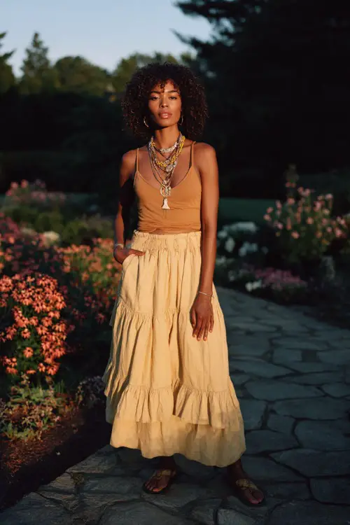 A Black woman wears a tiered boho midi skirt with a fitted top tucked in, paired with slip-on sandals and layered necklaces 