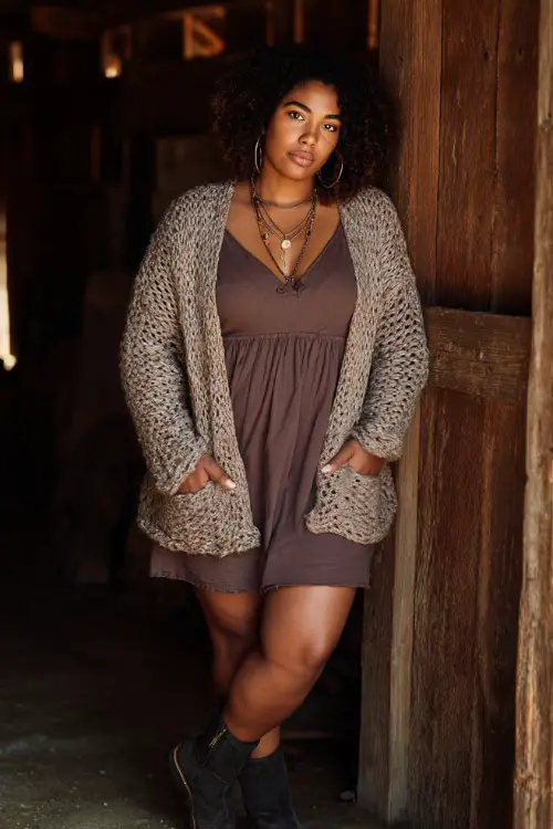 A curvy woman wears a crochet cardigan layered over a simple fitted dress, styled with ankle boots and bold jewelry, posing in a rustic outdoor setting with warm tones