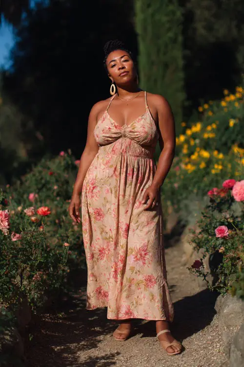 A curvy woman wears a flowy boho maxi dress with a defined waist and soft floral prints, paired with strappy sandals and statement earrings
