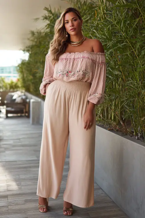 A plus-size woman wears a lightweight off-shoulder blouse with embroidery paired with high-waisted wide-leg pants 